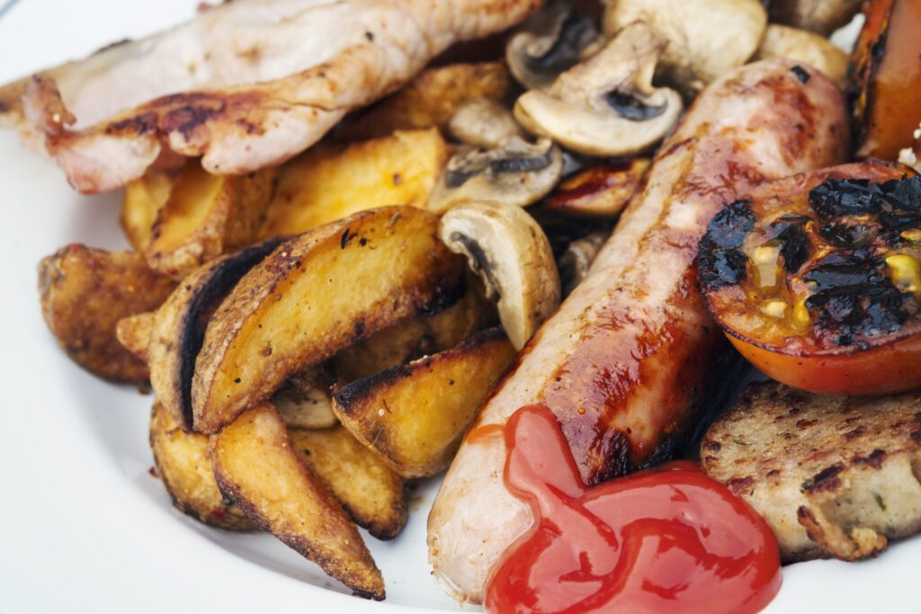 Close up of a plate of food, English Breakfast, bacon, mushrooms, sausage, grilled tomato, potatoes and tomato sauce.